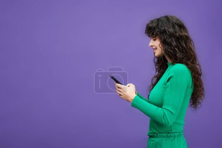 Téléchargez les photos : Vue latérale de la jeune femme brune en costume vert à l'aide d'un téléphone portable tout en envoyant des SMS ou en regardant des informations en ligne isolément - en image libre de droit