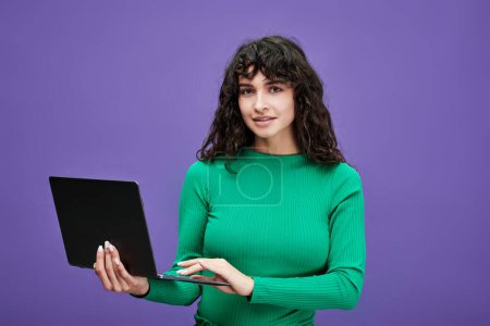 Foto de Joven profesor o estudiante seguro de sí mismo en jersey verde sosteniendo el ordenador portátil y la creación de redes mientras está de pie delante de la cámara en fondo violeta - Imagen libre de derechos