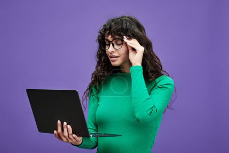 Téléchargez les photos : Jeune femme d'affaires ou professeur en pull vert et lunettes de vue regardant l'écran d'ordinateur portable tout en regardant à travers l'information en ligne - en image libre de droit