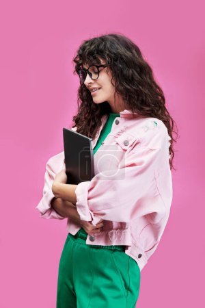 Téléchargez les photos : Vue latérale d'une jolie étudiante souriante portant des lunettes et des vêtements décontractés tenant un gadget mobile par la poitrine tout en se tenant debout sur fond rose - en image libre de droit