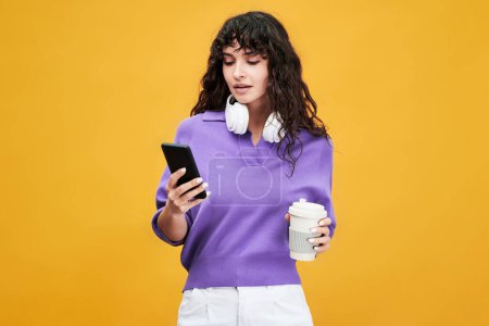 Téléchargez les photos : Étudiante mignonne en v-shirt lavande et en pantalon utilisant un téléphone portable et ayant une tasse de thé à la pause sur fond jaune - en image libre de droit