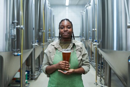 Téléchargez les photos : Portrait d'une travailleuse positive en uniforme montrant un verre de bière produit dans sa brasserie - en image libre de droit