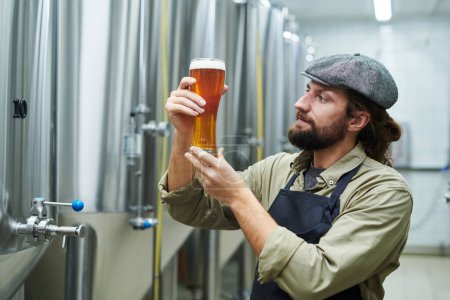 Téléchargez les photos : Ouvrier de brasserie positif en chapeau plat regardant un verre de bière avec une mousse épaisse - en image libre de droit
