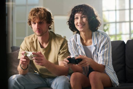 Téléchargez les photos : Joyeux jeune femme et son petit ami contrarié en tenue décontractée assis sur le canapé dans le salon à loisir et jouer à un jeu vidéo - en image libre de droit