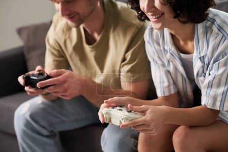Foto de Recortado disparo de joven pareja descansada disfrutando de un nuevo videojuego mientras están sentados uno al lado del otro y pulsando botones de joysticks - Imagen libre de derechos