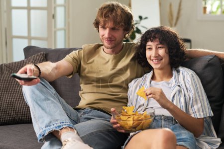 Téléchargez les photos : Joyeux jeune femme avec bol de croustilles assis sur le canapé à côté de son mari choisir quelque chose d'intéressant à regarder à la télévision - en image libre de droit