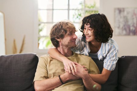 Téléchargez les photos : Joyeux jeune femme brune embrassant son mari assis sur le canapé et le regardant avec le sourire tout en passant le week-end à la maison - en image libre de droit