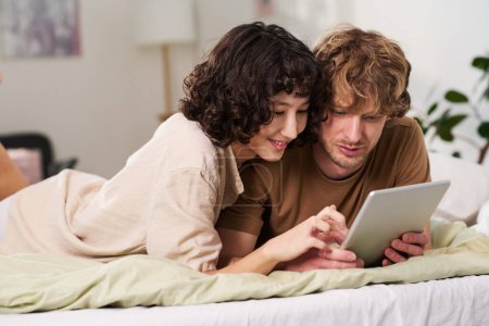 Photo for Young intercultural couple with tablet lying in bed after sleep in the morning and deciding what to order for breakfast from online menu - Royalty Free Image