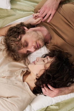 Téléchargez les photos : Plan ci-dessus de jeunes couples reposants les yeux fermés et les têtes tournées les unes vers les autres allongées sur le lit et dormant tôt le matin - en image libre de droit