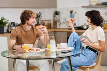 Téléchargez les photos : Joyeux jeune couple ayant des sandwichs aux légumes et du jus de fruits pour le petit déjeuner assis à table dans la cuisine et discutant des plans pour la journée - en image libre de droit