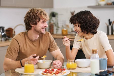 Foto de Feliz joven y su esposa comiendo copos de maíz con leche para el desayuno mientras está sentado junto a la mesa de la cocina servida con sándwiches y jugo - Imagen libre de derechos