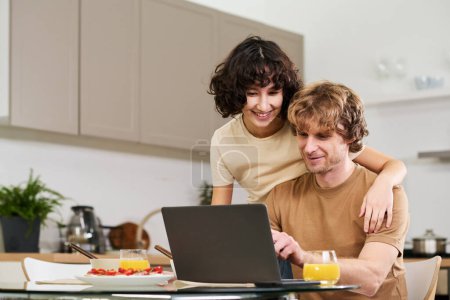 Photo for Happy young woman embracing her husband while both looking at laptop screen and choosing goods in online shop during breakfast - Royalty Free Image