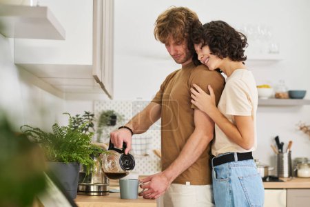 Photo for Young smiling woman standing close to her husband pouring coffee in grey mug and embracing him while looking forwards for breakfast - Royalty Free Image
