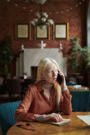 Téléchargez les photos : Jolie fille albinos avec smartphone par oreille communiquer avec un ami tout en étant assis à table avec un bloc-notes et passer ses loisirs dans un café - en image libre de droit