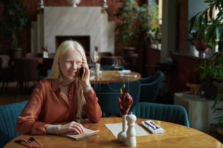 Téléchargez les photos : Jeune femme albinos gaie parlant à un ami sur un téléphone portable tout en étant assis à table dans un café confortable à loisir ou week-end et se reposer - en image libre de droit