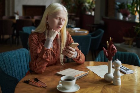 Téléchargez les photos : Reposé fille albinos en tenue décontractée intelligente regardant l'écran du smartphone tout en envoyant des SMS ou en faisant défiler les données en ligne tout en étant assis dans le café - en image libre de droit