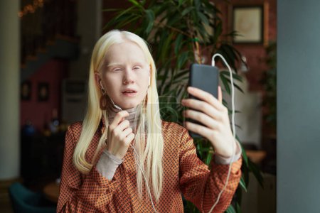 Photo for Youthful albino girl in earphones holding mobile phone in front of herself while looking at her friend on screen during communication in video chat - Royalty Free Image