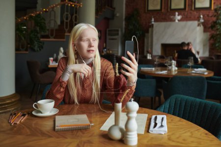 Photo for Albino girl with long hair looking at smartphone screen during communication with friend in video chat in cozy cafe - Royalty Free Image