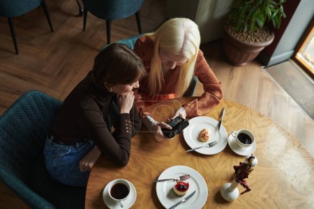Photo for Above angle of two young friendly women looking through photos in photocamera while sitting by table in cafe and having dessert - Royalty Free Image