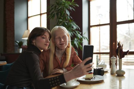 Foto de Dos chicas en ropa de casualwear inteligente mirando a la pantalla del teléfono inteligente mientras toma selfie por mesa en acogedora cafetería decorada con plantas verdes - Imagen libre de derechos