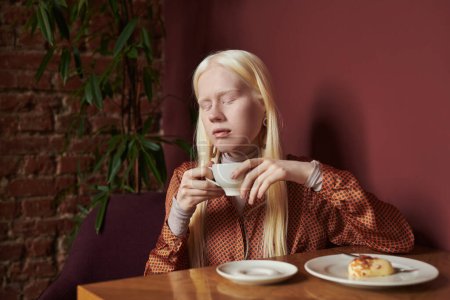 Téléchargez les photos : Jeune femme albinos sereine avec de longs cheveux blancs tenant une tasse de café tout en étant assis à table avec dessert dans un café avec des plantes vertes - en image libre de droit