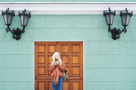 Téléchargez les photos : Serene fille albinos en jeans bleus et veste brune debout à côté de la porte d'entrée du bâtiment de théâtre décoré de lanternes classiques - en image libre de droit