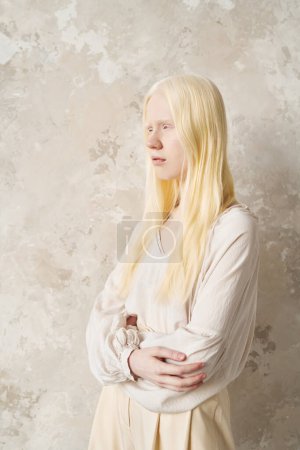 Foto de Mujer albina joven en ropa de casa de algodón blanco manteniendo sus brazos cruzados por el pecho mientras está de pie junto a la pared de mármol delante de la cámara - Imagen libre de derechos