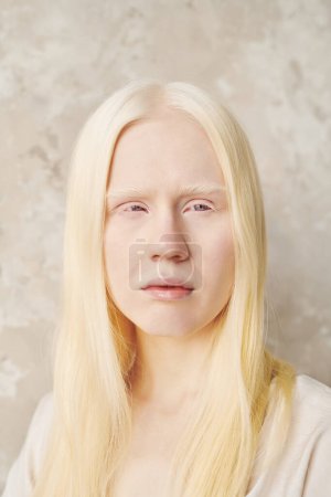 Photo for Adolescent albino girl with long smooth hair posing in front of camera during photo session while standing against white wall in isolation - Royalty Free Image