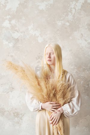Foto de Mujer albina joven con ramo de flores secas esponjosas en las manos manteniendo los ojos cerrados mientras está de pie junto a la pared delante de la cámara - Imagen libre de derechos