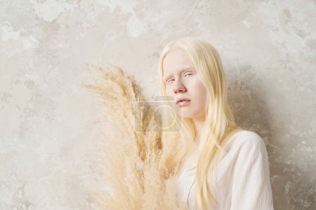 Foto de Retrato de belleza natural de chica albina pacífica sosteniendo ramo de flores secas mientras está de pie cerca de la pared blanca delante de la cámara - Imagen libre de derechos