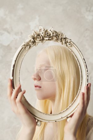Photo for Side view of young serene albino woman holding antique oval frame decorated with roses while posing in front of camera in studio - Royalty Free Image