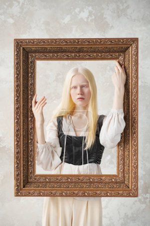 Photo for Cute albino girl in medieval attire keeping hands on large picture frame while standing behind it and posing during photo session in studio - Royalty Free Image