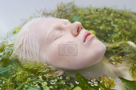 Photo for Close-up of pale face of albino girl lying in bath with warm pure water and green plants during relaxation and hygiene procedure - Royalty Free Image