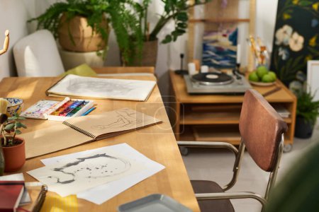 Téléchargez les photos : Papiers et bloc-notes avec croquis dessinés par l'artiste en home studio avec table, chaise, plantes vertes, aquarelles, tourne-disques et autres choses - en image libre de droit
