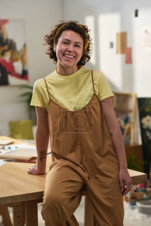 Téléchargez les photos : Jeune artiste féminine réussie en tenue décontractée regardant la caméra tout en se tenant près de la table en bois pendant le travail en studio à la maison - en image libre de droit