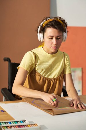 Téléchargez les photos : Jeune artisan sérieux handicapé écoutant de la musique dans des écouteurs et dessinant avec des crayons sur une feuille de papier - en image libre de droit