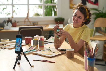 Junge lächelnde kreative Frau mit Pinsel und Gouache sitzt am Arbeitsplatz und schaut während des Meisterkurses in die Smartphone-Kamera