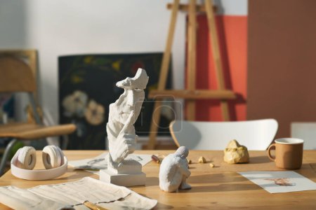 Téléchargez les photos : Lieu de travail de l'artisan moderne avec des sculptures d'argile et de gypse, croquis sur papier, tasse et écouteurs contre la peinture et le chevalet - en image libre de droit