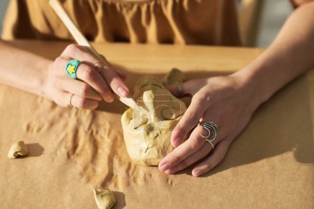 Téléchargez les photos : Mains de jeune femme créative avec outil en bois coupant les yeux sur le visage de la figurine humaine en argile tout en étant assis sur le lieu de travail dans le home studio - en image libre de droit