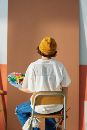 Téléchargez les photos : Vue arrière de la jeune artiste féminine en tenue décontractée assise devant une pièce vierge préparée pour peindre un futur chef-d'œuvre - en image libre de droit