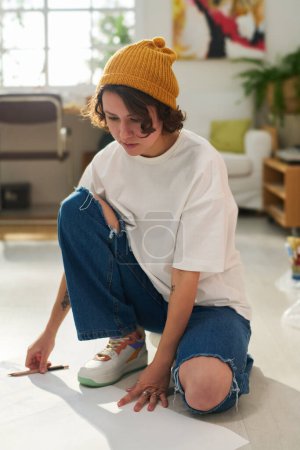 Téléchargez les photos : Jeune artiste féminine coûteuse penser à un nouveau sujet pour les œuvres d'art tout en étant assis sur le sol parmi une grande feuille de papier dans le home studio - en image libre de droit