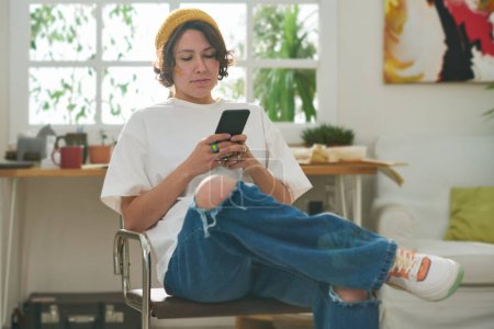 Foto de Mujer seria joven en ropa casual elegante sentado en la silla en el estudio en casa y el uso de teléfono móvil para ver vídeo en línea - Imagen libre de derechos