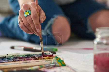 Téléchargez les photos : Focus sur la main de la jeune femme créative avec bague bijou mettre pinceau en aquarelle tout en étant assis sur le sol de l'atelier et la création d'œuvres d'art - en image libre de droit