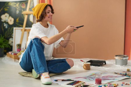 Téléchargez les photos : Joyeux jeune peintre créative avec smartphone assis sur le sol et prenant des photos de nouvelles œuvres peintes à l'aquarelle sur papier - en image libre de droit