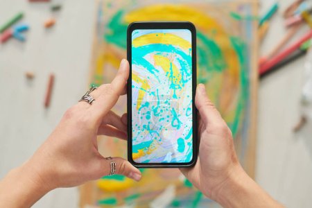 Téléchargez les photos : Au-dessus de l'angle du smartphone tenu par la jeune créative peintre femme prenant des photos de nouvelles œuvres abstraites peintes avec des aquarelles bleues et jaunes - en image libre de droit