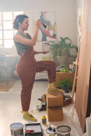 Foto de Vista lateral de la joven morena artista femenina con pequeño cubo y pincel de pie delante de un gran panel mientras se crea una nueva obra maestra - Imagen libre de derechos