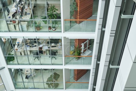 Photo for Part of modern multi-storey building with rows of desks, armchairs and green plants in openspace offices behind large windows - Royalty Free Image