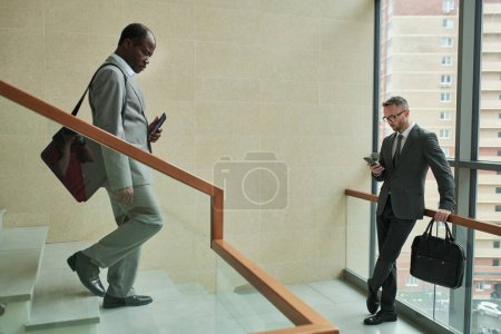 Photo for Mature African American businessman walking downstairs while young male entrepreneur using smartphone inside modern business center - Royalty Free Image