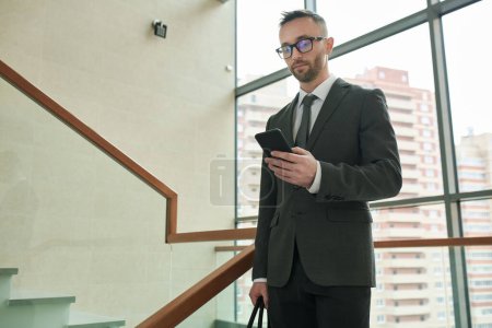 Photo for Young chief executive office in elegant suit and eyeglasses using mobile phone while standing inside business center between staircases - Royalty Free Image