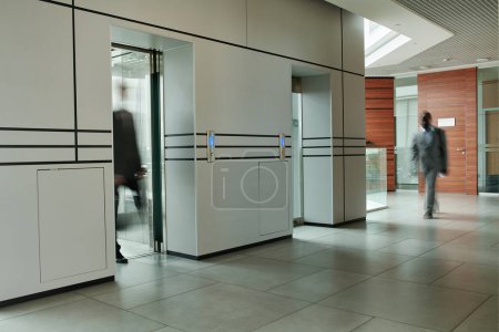Foto de Movimiento borroso de dos empleados masculinos en el espacioso pasillo del moderno centro de negocios mientras uno de ellos sale del ascensor - Imagen libre de derechos